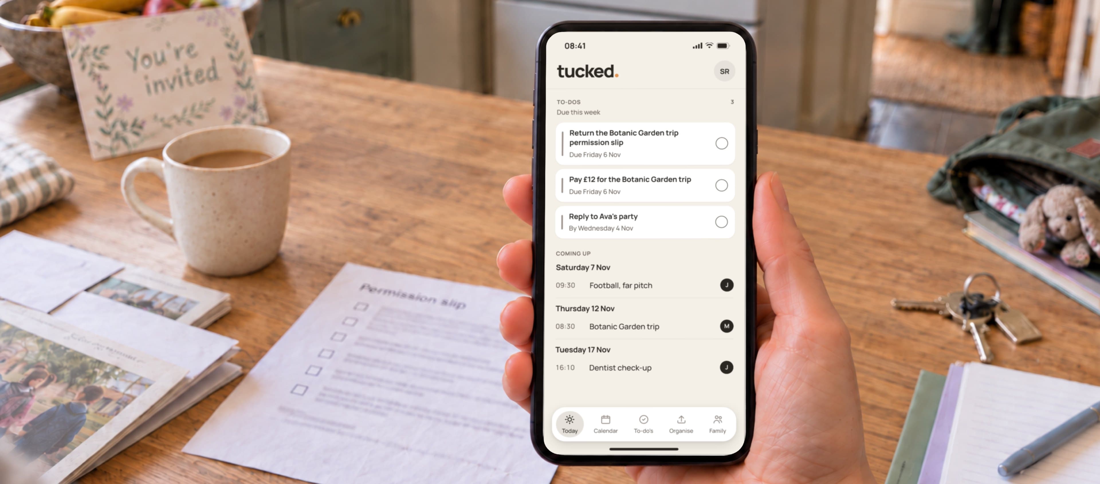 A phone showing the tucked Today screen, with this week's to-dos and dates, in front of family paperwork on a kitchen table.